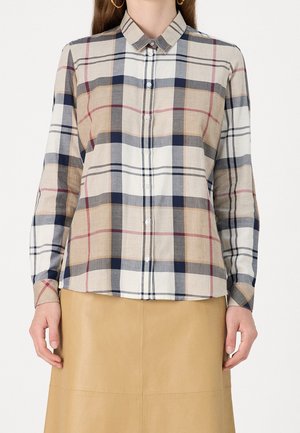 Vrouw draagt een overhemd met beige, marineblauw, rood en wit geruit patroon met lange mouwen en een beige rok, staande tegen een effen achtergrond.