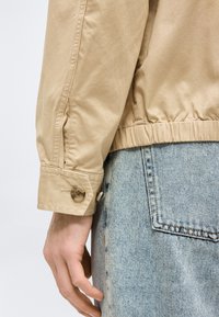 Μανίκι μακρυμάνικου μπεζ σακακιού με λουράκι και κουμπί, ελαστική μέση, και τσέπη ανοιχτού μπλε τζιν δίπλα σε χαλαρό χέρι.