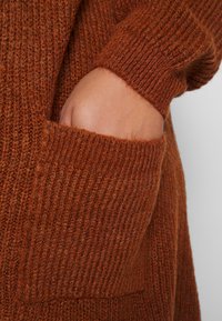 Maglione lavorato a maglia marrone con una texture a coste e una tasca. La manica si estende fino al polso, mostrando una vestibilità aderente.