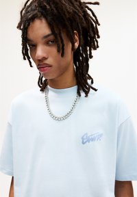 Lyseblå oversized t-shirt med en blød tekstur, der har et diskret grafisk logo i en mørkere blå farve på venstre bryst. Bæres med en kraftig sølvkæde.