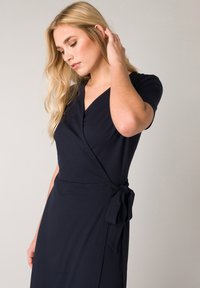 Navyblaues Wickelkleid aus weichem Stoff mit kurzen Ärmeln und gebundenem Taillengürtel. Es hat einen V-Ausschnitt und eine glatte Textur mit minimalen Details.