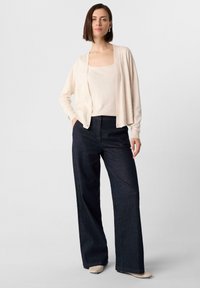 Beige cardigan over een lichtbeige top, gecombineerd met donkerblauwe wijde broek. De outfit heeft een ontspannen pasvorm en zachte texturen.