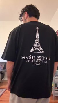 Man with curly hair facing away wearing a black shirt with a white Eiffel Tower graphic and reversed French text.