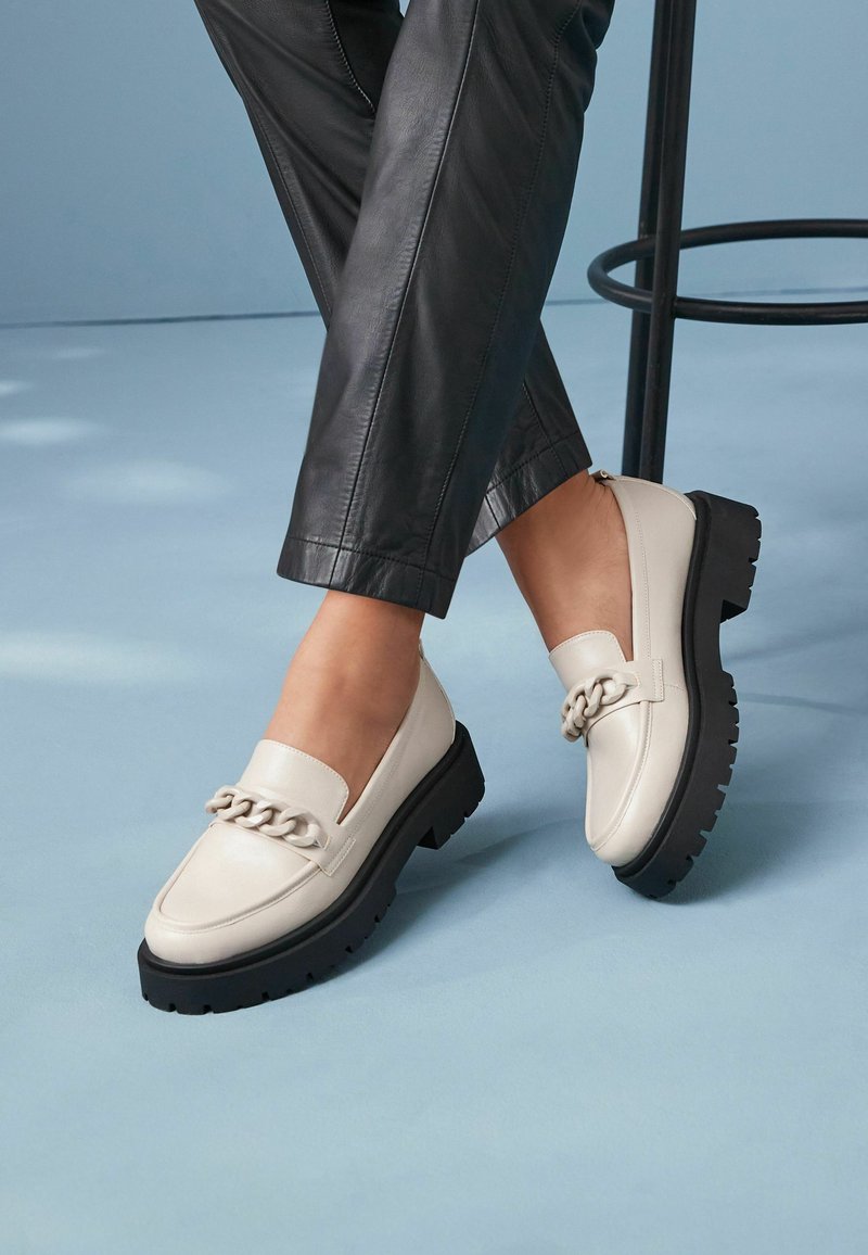 Vaaleanbeige loafers, joissa on paksu musta platform-pohja ja koristeellinen ketju kengän päällä. Kengässä on sileä nahkapinta.