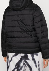 Black puffer jacket with horizontal quilted pattern, a hood, and a smooth, shiny texture. Worn over white tie-dye pants.