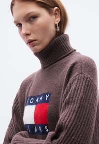 Σκούρο καφέ πουλόβερ με λαιμόκοψη ζιβάγκο από μαλακό μάλλινο ύφασμα με ανάγλυφη υφή και εμφανές κεντημένο λογότυπο σε κόκκινους, λευκούς και μπλε χρωματισμούς.