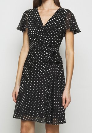 Kobieta w czarnej sukience kopertowej w białe kropki, z prześwitującymi, zwiewnymi rękawami i wiązaniem z boku w talii.