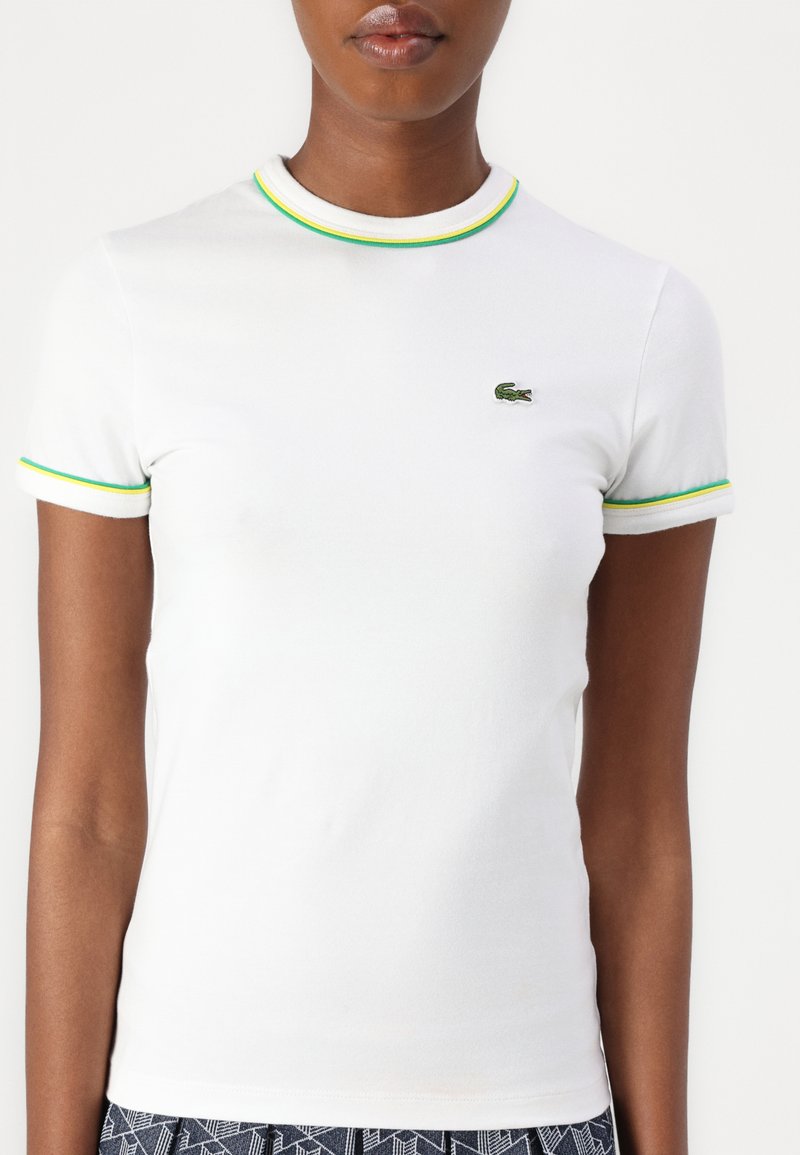 Biała koszulka z krótkim rękawem z zielonymi i żółtymi detalami. Posiada małe, haftowane zielone logo krokodyla na piersi. Materiał bawełniany.