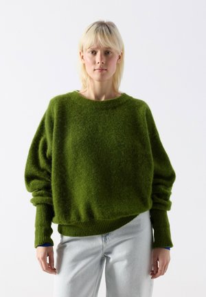 Femme blonde portant un pull vert ample et duveteux et un jean bleu clair, debout devant un fond blanc uni.