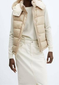 Beige gesteppte Weste mit abnehmbarem Kapuzen, getragen über einem hellbeigen Pullover und einem neutralen Rock. Verfügt über Druckknöpfe und seitliche Taschen.