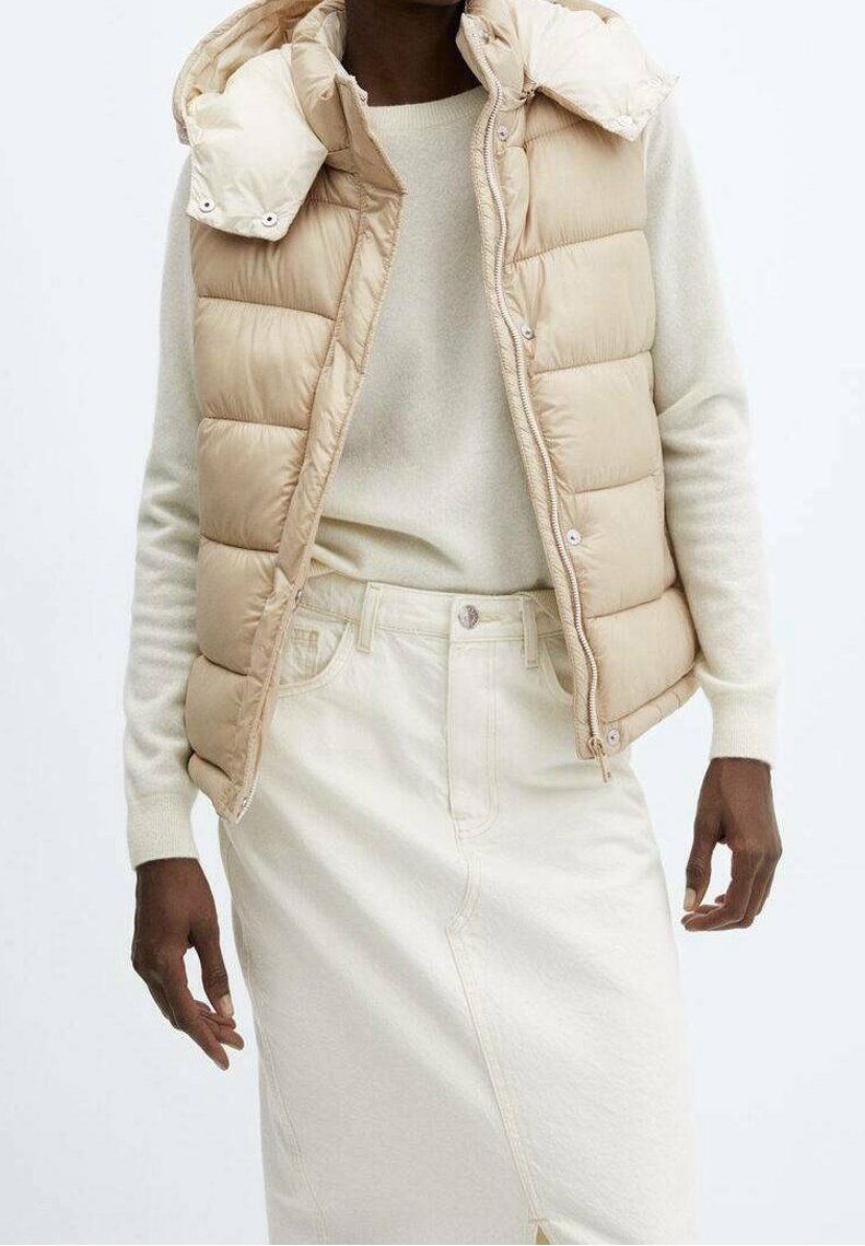 Beige gesteppte Weste mit abnehmbarem Kapuzen, getragen über einem hellbeigen Pullover und einem neutralen Rock. Verfügt über Druckknöpfe und seitliche Taschen.