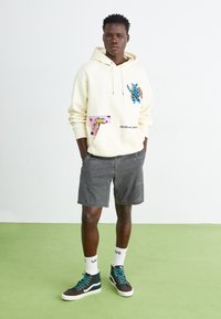 Cream hoodie with colorful graphic patches, grey shorts, and black high-top sneakers with teal laces, standing on green carpet.