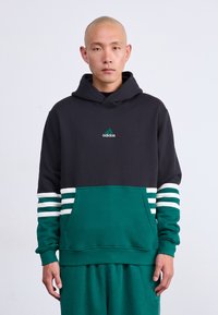 Moški z britvasto glavo, oblečen v črn in zelen Adidas kapuco z belimi črtami na rokavih in usklajenih zelenih hlačah, stoji ob belem ozadju.