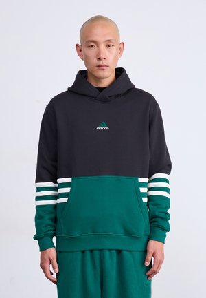 Bărbat cu cap ras, îmbrăcat într-o hanorac Adidas negru și verde, cu dungi albe pe mâneci și pantaloni verzi asortați, stând în fața unei fundal alb.