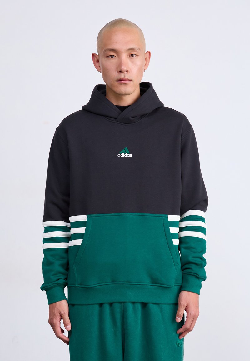 Hombre con la cabeza rapada que lleva una sudadera negra y verde de Adidas con franjas blancas en las mangas y pantalones verdes a juego, de pie contra un fondo blanco.
