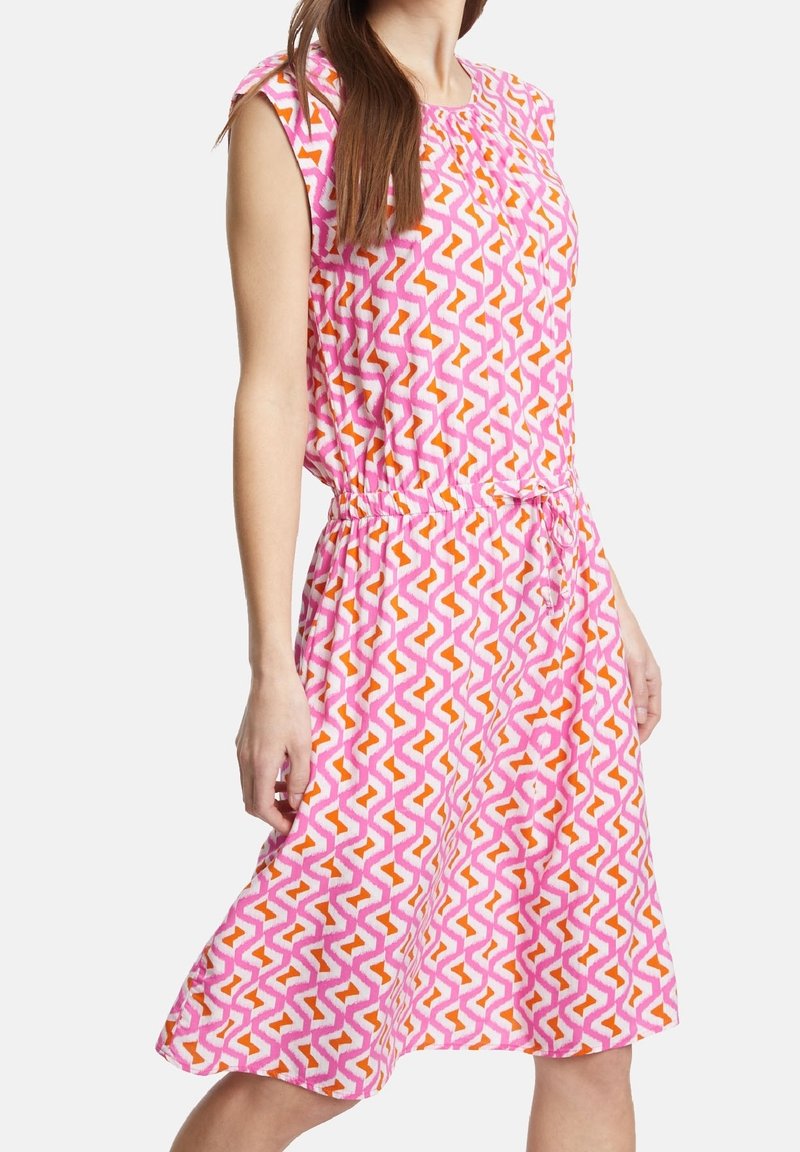 Robe rose avec un design à motifs présentant des formes en zigzag orange et blanches. Tissu léger, manches courtes et taille élastique.