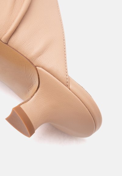 Μπεζ δερμάτινο παπούτσι με χαμηλό τακούνι, ορατό από πίσω, με στρογγυλεμένο τακούνι και υφή δερμάτινης επιφάνειας.