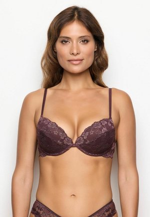 Donna con capelli ondulati che indossa un set coordinato di reggiseno e mutandine in pizzo viola, in piedi su uno sfondo bianco semplice, rivolta di fronte.