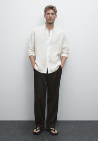 Homme portant une chemise blanche à boutons avec des manches retroussées, un pantalon noir ample et des sandales noires, se tenant contre un mur gris uni.