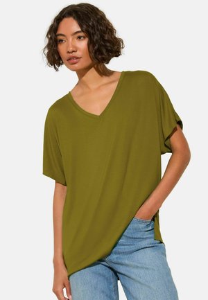 Kobieta o kręconych włosach, ubrana w oliwkowozielony top z krótkim rękawem i dekoltem w serek oraz jasnoniebieskie dżinsy, z jedną ręką w kieszeni, na jednolitym tle.