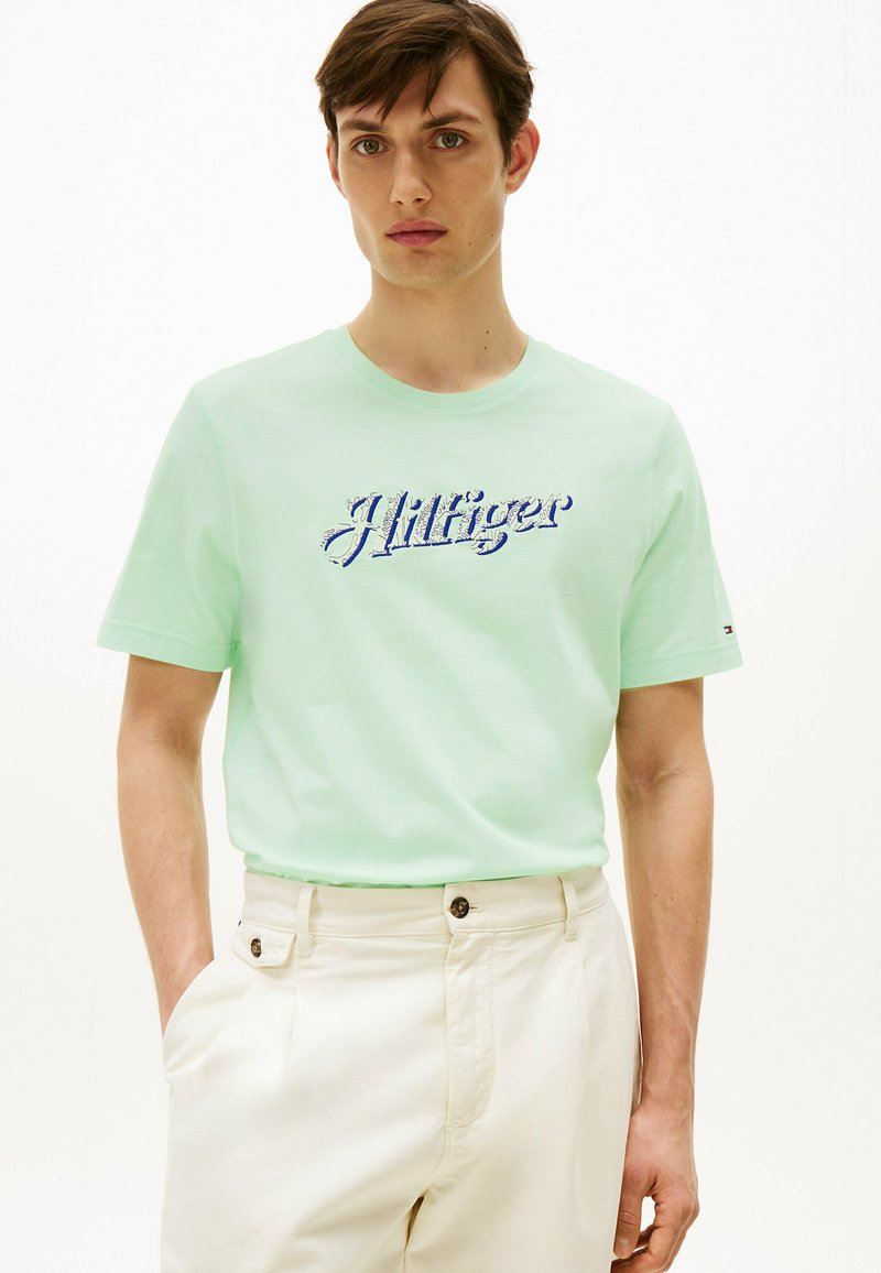 Camiseta de algodón color verde menta con "Hilfiger" bordado en azul. Manga corta, cuello redondo, combinada con pantalones color crema.