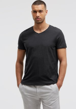 Mann mit kurzen Haaren, der ein eng anliegendes schwarzes V-Ausschnitt-T-Shirt und hellgraue Hosen trägt, steht mit den Händen in den Taschen vor einem schlichten Hintergrund.