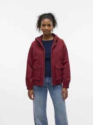 Veste légère rouge avec capuche, poignets élastiques et deux poches avant. Portée par-dessus un pull bleu foncé à texture côtelée.