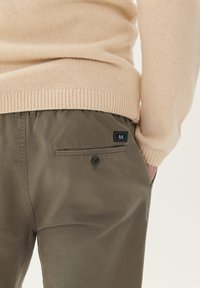 Pulover tricotat bej, asortat cu pantaloni verde măsliniu care au un buzunar la spate și detalii cu buton. Textură netedă, croială confortabilă.