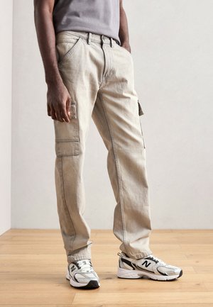 Pantalons cargo beige en coton, avec poches latérales et coupe décontractée. Portés avec des baskets blanches et noires sur un sol en bois.
