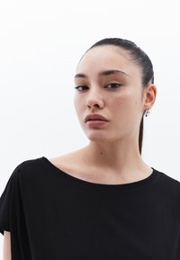 Musta, löysä t-paita, jossa on leveä kaula-aukko ja pehmeä tekstuuri. Mallilla on taakse kammattu hiustyyli ja vähäiset asusteet, tausta on selkeä.