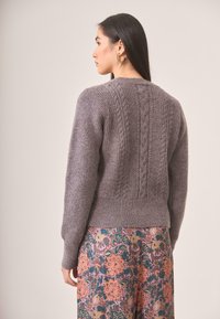 Suéter gris de punto con un patrón de trenzado texturizado y puños acanalados, llevado sobre una falda con estampado floral, visto desde la parte posterior.