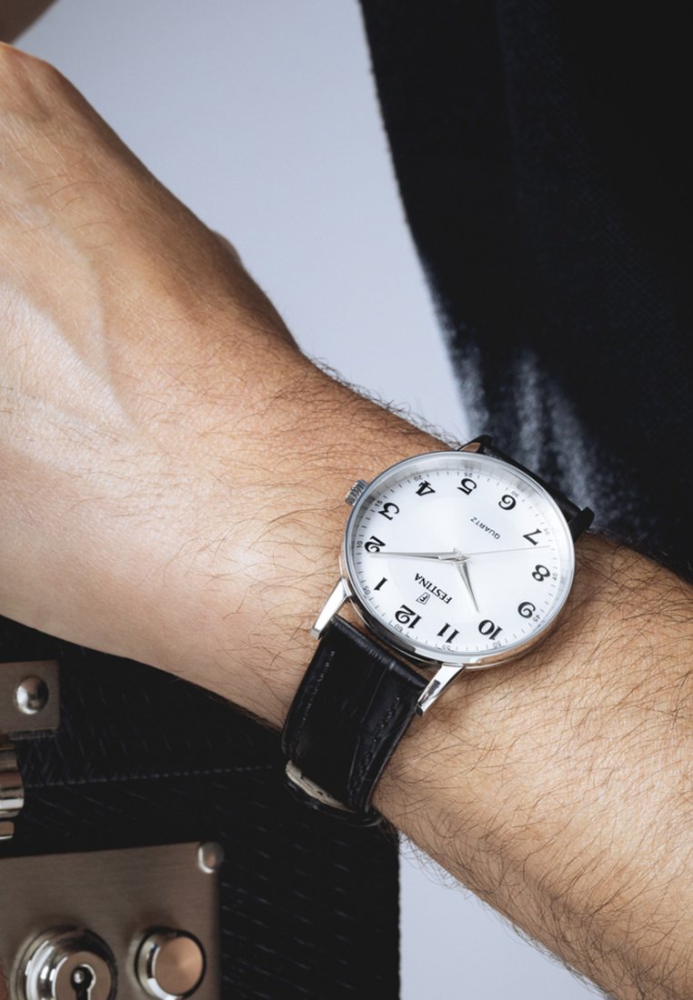 Poignet avec une montre à bracelet en cuir noir affichant 11h15, tenant une mallette noire texturée avec une serrure argentée.