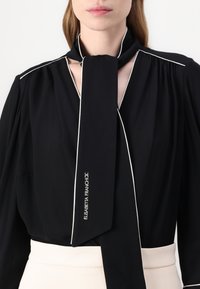 Črna bluzica z ovijalnim trakom, ki ima bele obrobe ob zapenjanju in ovratniku. Na traku je logo "Elisabetta Franchi".