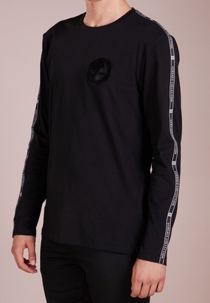 Mies, joka pukeutunut mustaan pitkähihainen paitaan, jossa on Versace Collectionin logo rinnassa ja merkkiraidat molemmissa hihoissa.