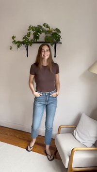 Camiseta ajustada marrón, jeans recortados azul claro y chanclas negras. Entorno interior con una estantería que sostiene una planta verde arriba. Suelo de madera, silla clara.