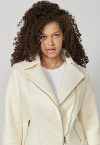 Krém színű műbőr dzseki texturált felülettel, puha fehér gallérral és ezüst cipzárakkal az elején és a zsebeknél.