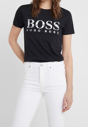 Kvinde iført en sort Hugo Boss T-shirt med hvidt logo og hvide bukser med høj talje, stående foran en ensfarvet baggrund.