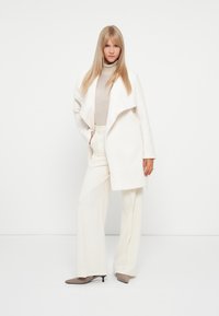 White, structured coat with a draped collar over a light beige turtleneck and wide-legged white pants, paired with gray pointed heels.