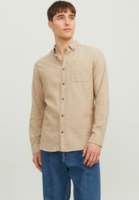 Beige Langarmhemd mit Button-Down-Kragen, einer einzelnen Brusttasche und braunen Knöpfen, aus strukturiertem Stoff. Getragen mit blauen Jeans.