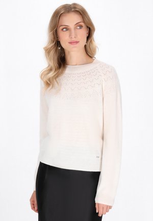 Helles, cremefarbenes Strickpullover mit strukturiertem Schößchen und Rautenmuster sowie gerippten Bündchen. Kombiniert mit einem schwarzen Rock.