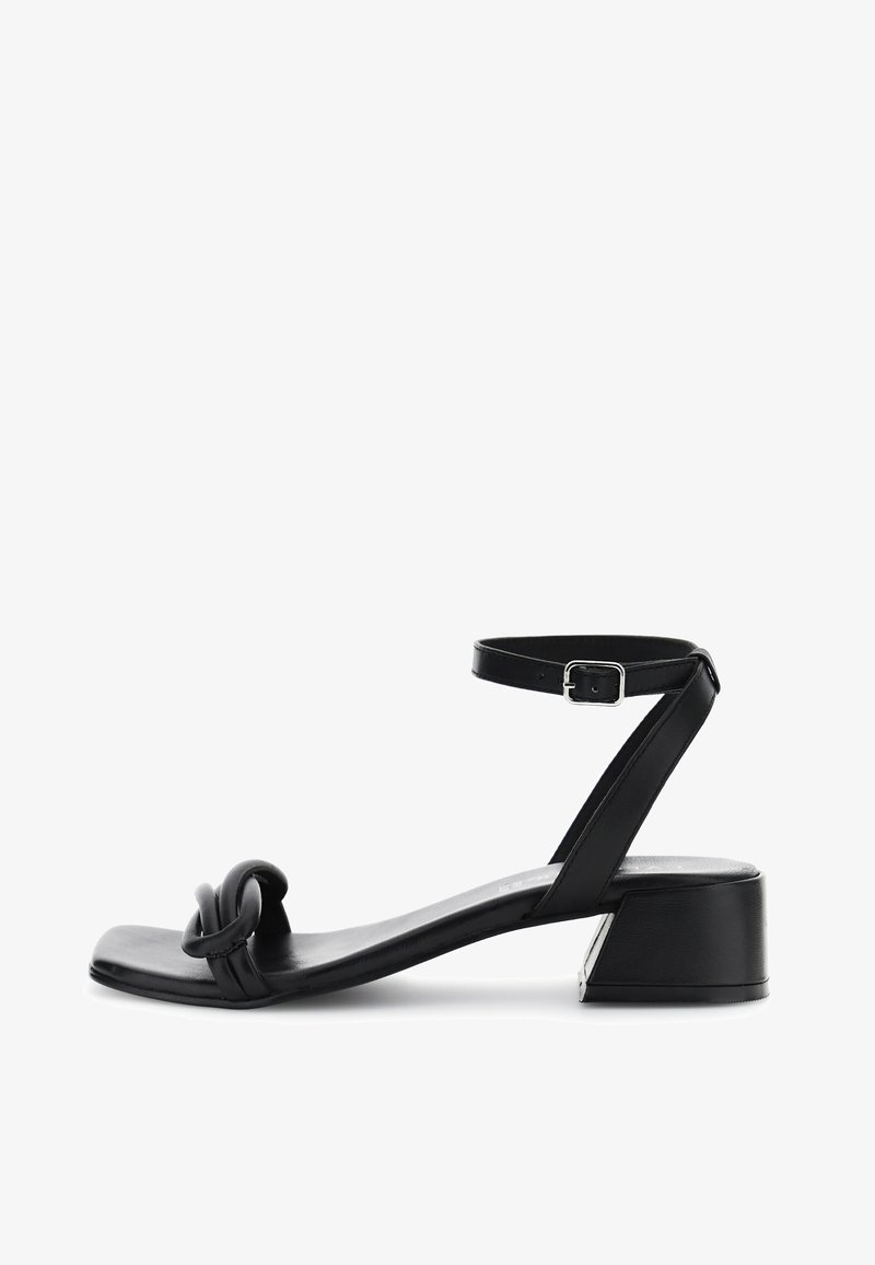 Musta sandaali matalalla lohkaremmillä, jossa on tasapohja, nilkkaremmi ja solmittu yksityiskohta varpaassa. Silkkinen nahkapinta.