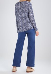 Woman wearing a long-sleeve floral blouse, straight-leg blue jeans, and beige loafers, standing facing away against a plain background.