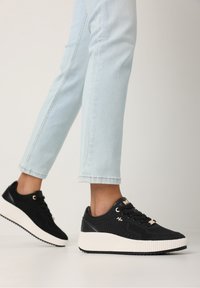 Svarta texturerade sneakers med vit gummisula och guldiga inslag, med snörning och syntetmaterial. Jeansen är synliga.