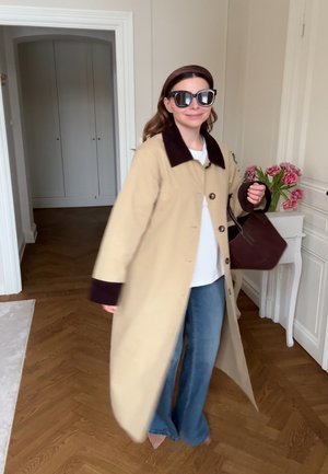 Frau trägt einen beigen Mantel mit dunklem Kragen und Manschetten, blaue Jeans, Sonnenbrille, trägt eine dunkelbraune Handtasche in einem Raum mit Holzboden und rosa Blumen.
