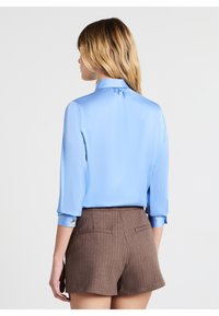 Blusa di raso azzurro chiaro con maniche lunghe e colletto alla francese, abbinata a pantaloni corti marroni a righe sottili. La blusa ha i polsini arrotolati.