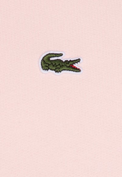 Un tissu en maille rose présente un logo de crocodile brodé en vert avec des détails rouges, bordé de coutures blanches.