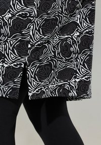 Der schwarz-weiße Stoff mit Mustern zeigt Kurven und kantige Designs. Der Saum des Kleidungsstücks hat einen geraden Schnitt und wird mit schwarzen Leggings kombiniert.