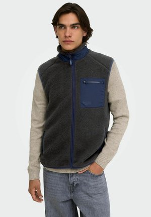 Jeune homme portant un gilet polaire gris foncé zippé avec une poche bleu marine et des manches en tricot beige, debout avec une main dans la poche du jean.