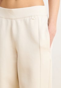 Svetlo bež hlače s visokim pasom, z gladko in mehko teksturo, opremljene s stranskimi žepi in poudarjeno zanko na pasu.