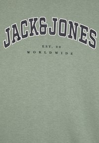 Zöld pamut pulóver, amelyen a "JACK&JONES" felirat vastag fekete betűkkel szerepel, alatta pedig kisebb betűmérettel az "EST. 90" és a "WORLDWIDE" felirat található.